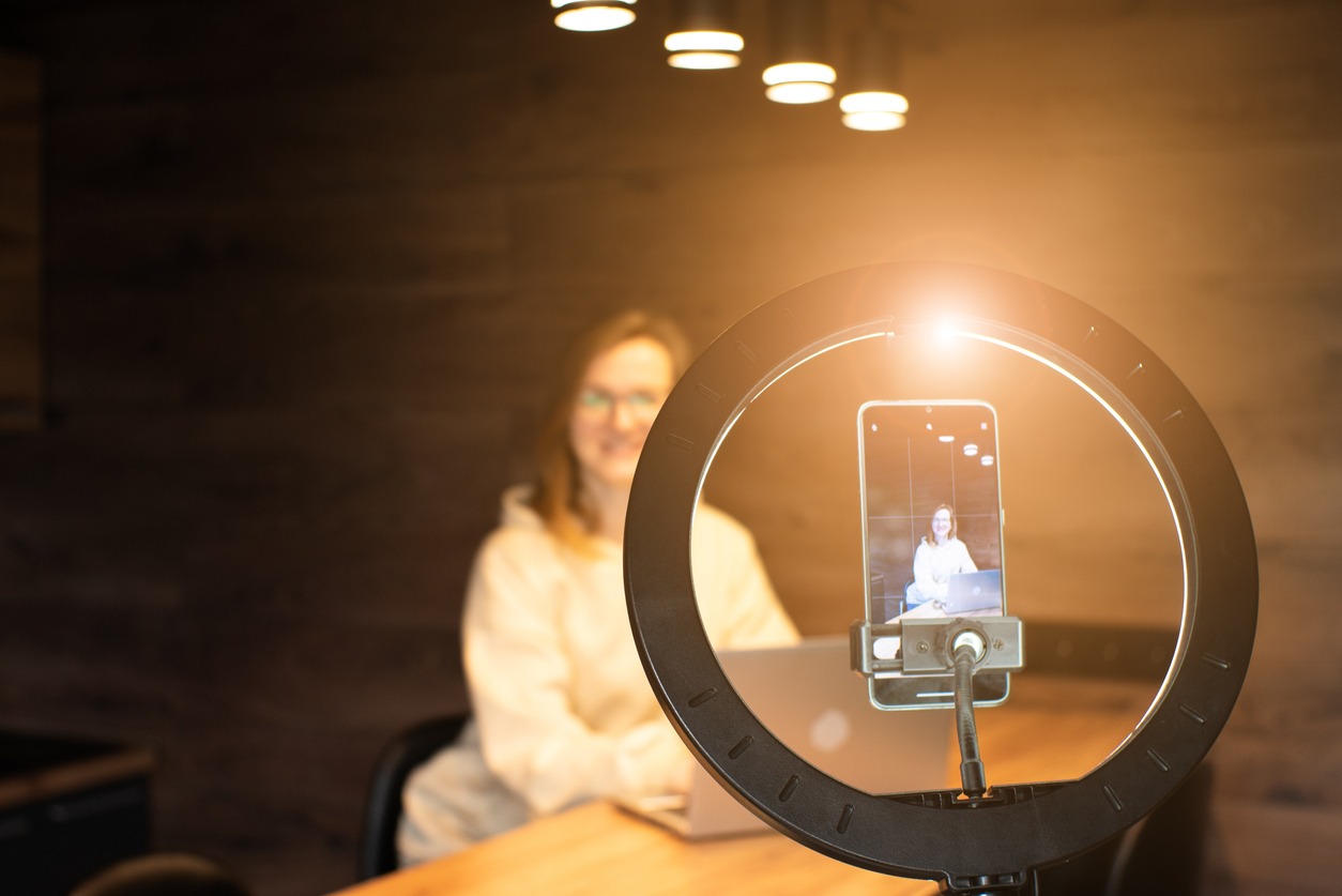Social media influencer creating content at modern studio sharing online stream using smartphone and laptop. Girl blogger smiling and speaking recording video with ring lamp. Online training courses.